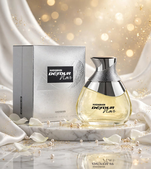 Al Haramain Detour Noir perfume bottle and its metallic silver box, displayed on a marble pedestal with white satin drapery and gold bokeh in the background.